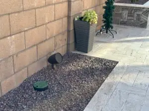 A neatly arranged backyard corner with a brick wall, a large, gray rectangular planter with green and yellow leafy plants, a small cone-shaped spotlight on the ground, and a small green sprinkler valve cover surrounded by gravel. A potted plant stands on tiled pavement.