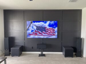 A modern home entertainment setup features a wall-mounted flat-screen TV displaying an American flag waving against a blue sky with clouds. The dark panelled wall serves as a backdrop, flanked by black speakers and subwoofers on either side.