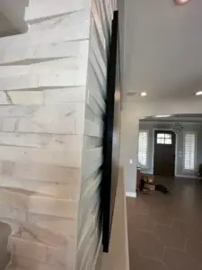 A side view of a thin, wall-mounted flat-screen TV in a living room with a textured white stone wall. The background shows the entryway with an open wooden door, a chandelier, and large floor tiles. Some boxes and cables are visible on the floor.