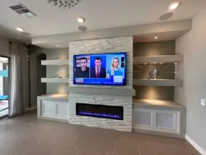 A modern living room features a wall-mounted flat-screen TV displaying a news broadcast. The TV is set above a contemporary electric fireplace. The wall is adorned with minimalist shelves holding decorative pieces, and the room has a neutral color palette.