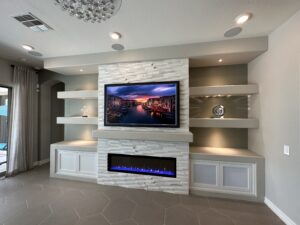 A modern living room features a TV mounted above an electric fireplace with a stone surround. Recessed shelves on either side hold decorative items, while cabinets below provide additional storage. Tile flooring and neutral colors complete the elegant look.