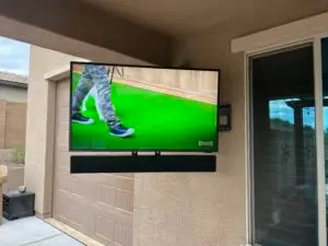 A flat-screen TV showing a person wearing camouflage pants and black sneakers walking on a green surface. The TV is mounted outdoors on a beige wall near a sliding glass door and window with closed blinds. There is a partial view of a garage and some outdoor items.